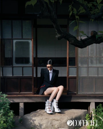 Fashion editorial photo: An adult woman in a black suit and white platform shoes sits on a stone step outside a traditional Japanese house.
