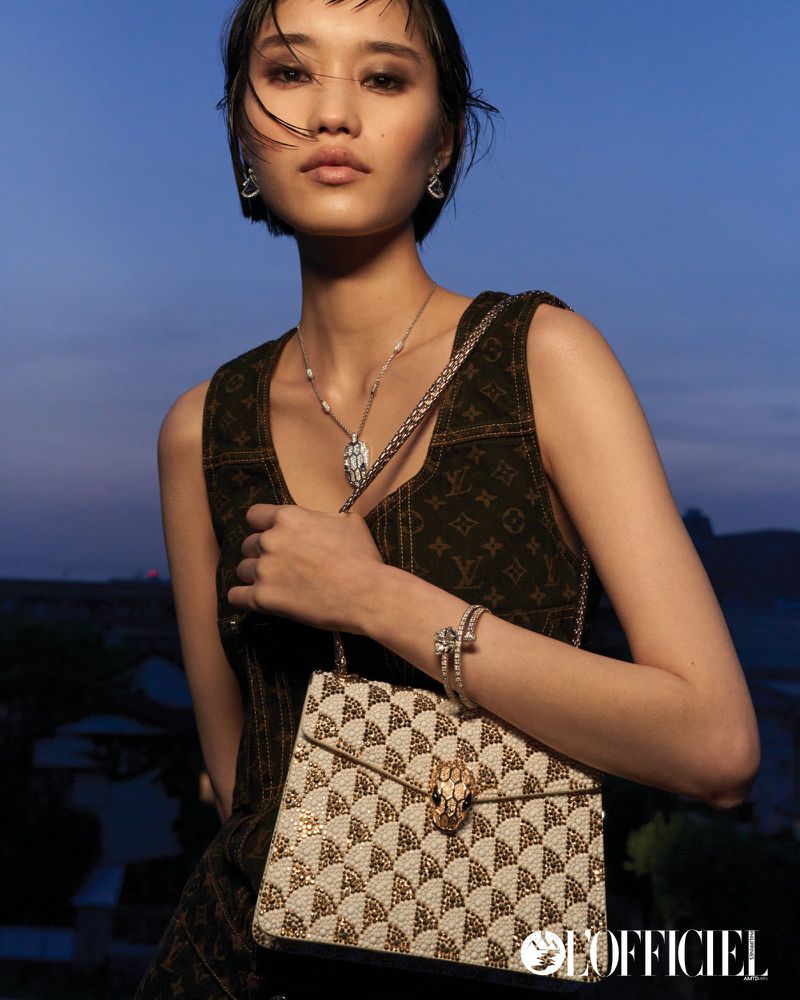 An Asian woman models jewelry and a patterned handbag in an outdoor setting with a blurred urban background at dusk or dawn.