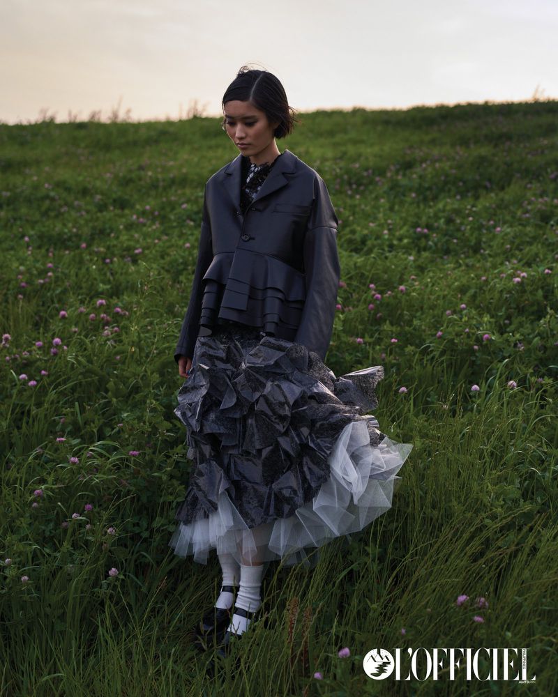 Young woman in dramatic dark fashion poses in a grassy field under a cloudy sky, wearing a jacket and ruffled skirt.