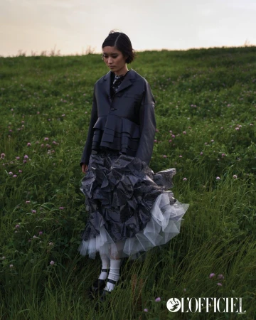 Young woman in dramatic dark fashion poses in a grassy field under a cloudy sky, wearing a jacket and ruffled skirt.