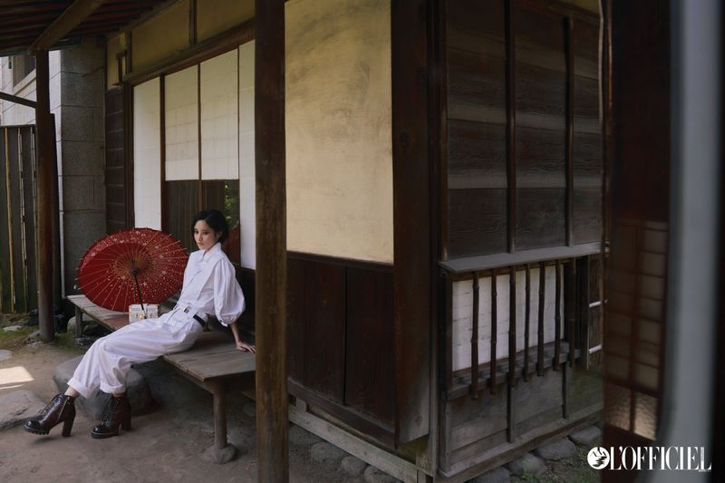 A woman in a white jumpsuit sits on a wooden bench in front of a traditional Japanese building, an umbrella rests on the bench.