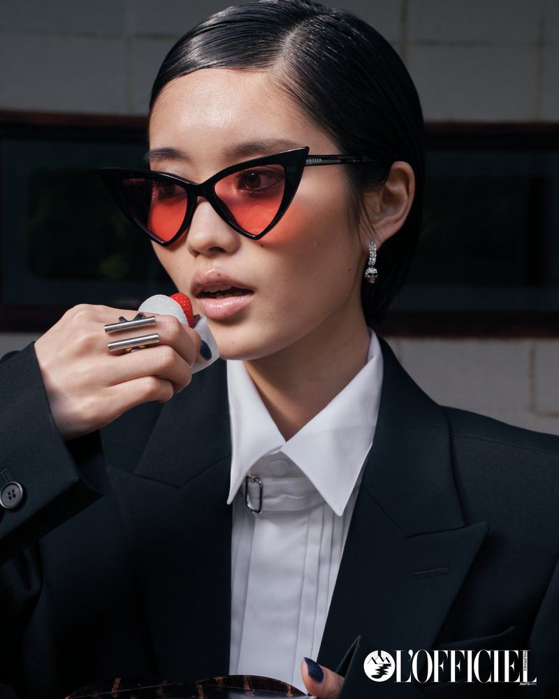 Stylish woman wearing sunglasses and suit jacket, posing indoors while holding a white object. L'Officiel logo in lower-right corner.