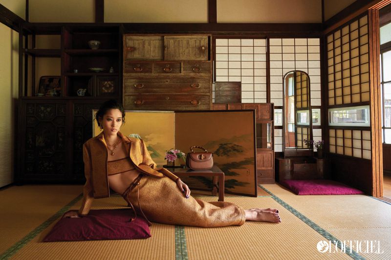 Adult woman in a brown two-piece outfit reclining on a maroon cushion, indoors on a tatami mat floor with brown antique furniture.