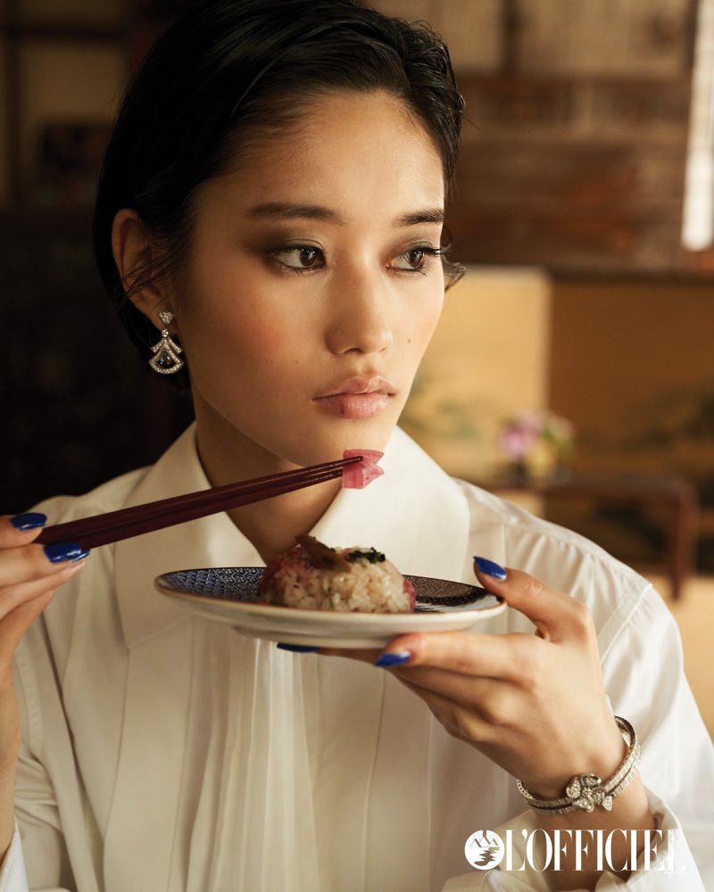 Asian woman with elegant jewelry holds a plate of sushi with chopsticks in a fashion editorial with a blurred background.