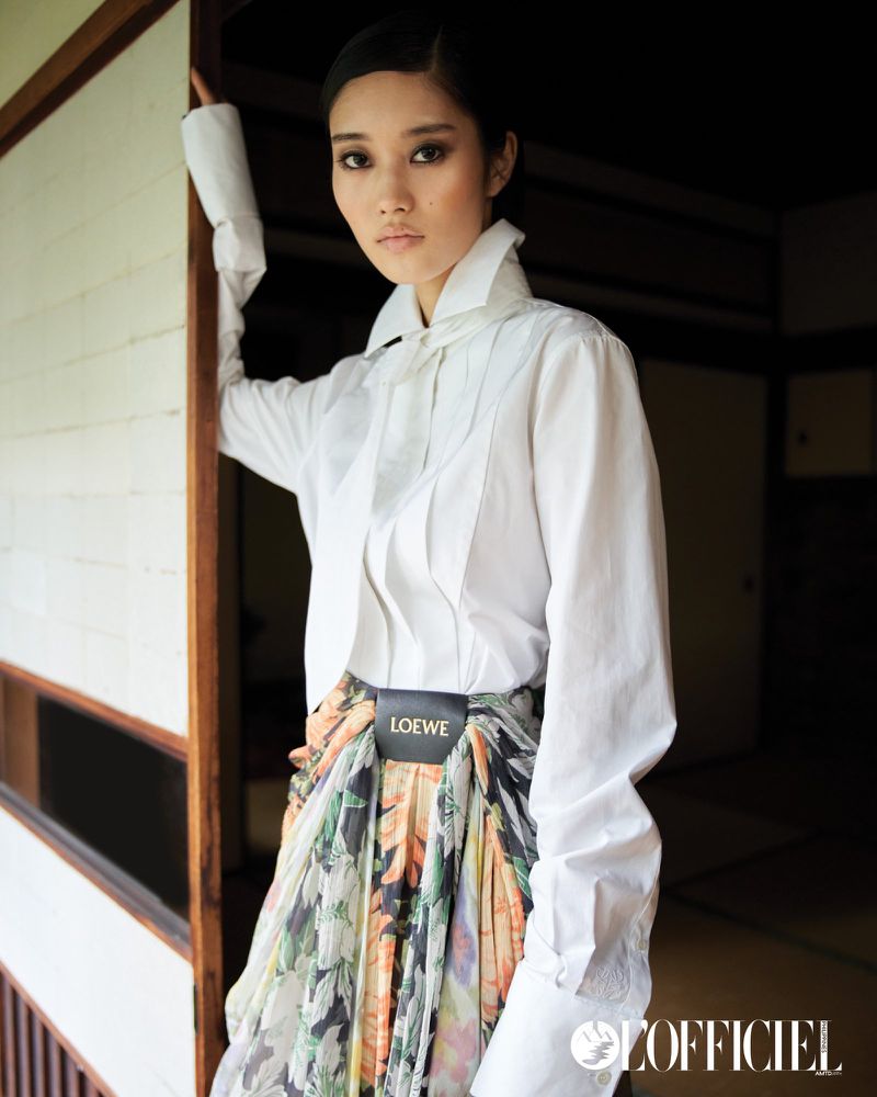 Indoor portrait of a young Asian woman in a white collared shirt and floral skirt, posing near a doorway, with a magazine logo.