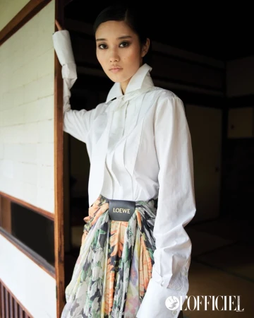 Indoor portrait of a young Asian woman in a white collared shirt and floral skirt, posing near a doorway, with a magazine logo.