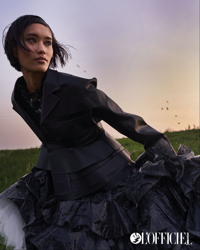 A woman in a black dress and jacket poses in a grassy field against a soft, light sky. The fashion editorial features natural lighting.