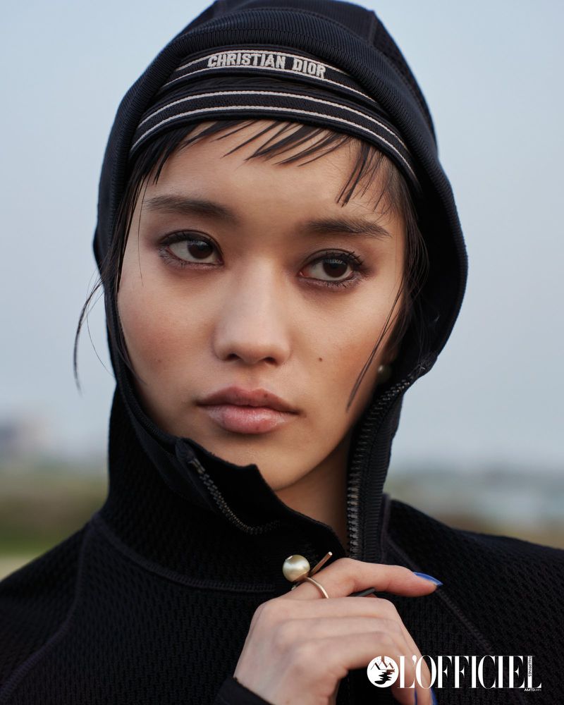 Close-up portrait of a young woman with an open black hooded jacket and Christian Dior branded headscarf, posing in a soft, outdoor light.