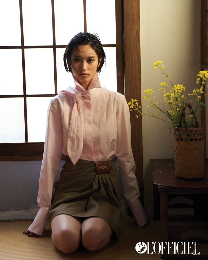 A brunette woman kneels wearing a pink blouse, olive skirt, and a thoughtful expression. She is indoors near a window with natural light.