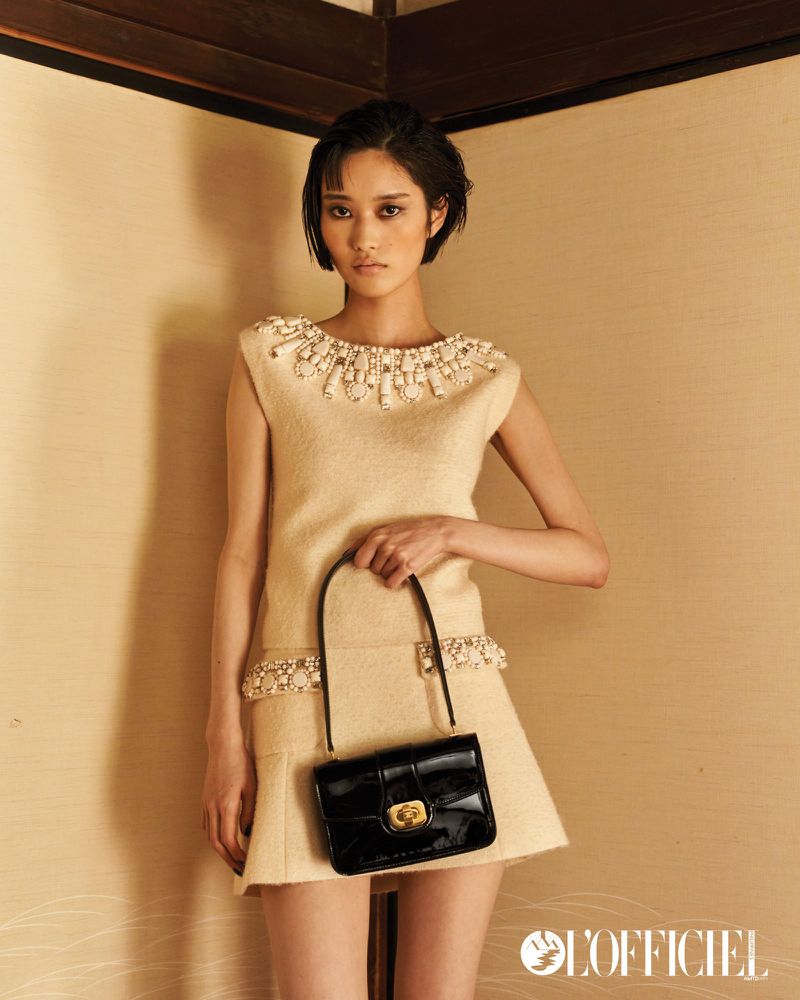 Asian woman in a sleeveless dress with pearl embellishments holding a black purse in a studio with neutral colored walls.