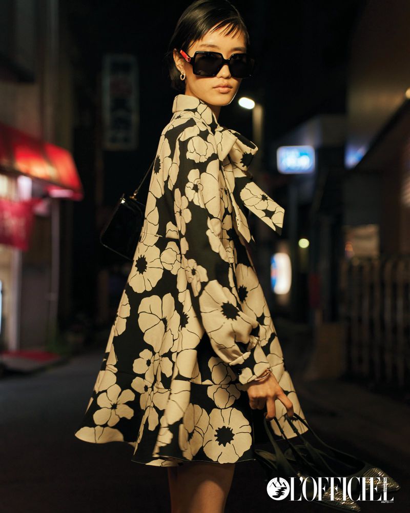 Woman in a floral print coat, sunglasses, and a black handbag standing on a city street at night illuminated by streetlights.