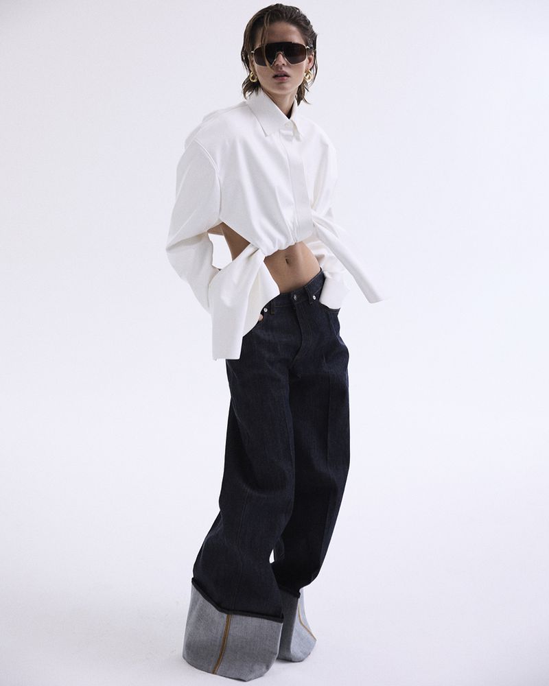 A woman poses in an open white shirt and wide-leg dark jeans against a white background, showcasing a casual yet fashionable look.