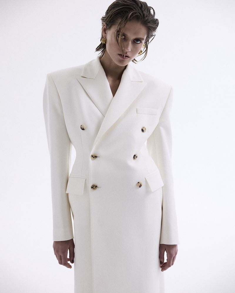 A female model in a white double-breasted coat with broad shoulders poses against a stark white background.