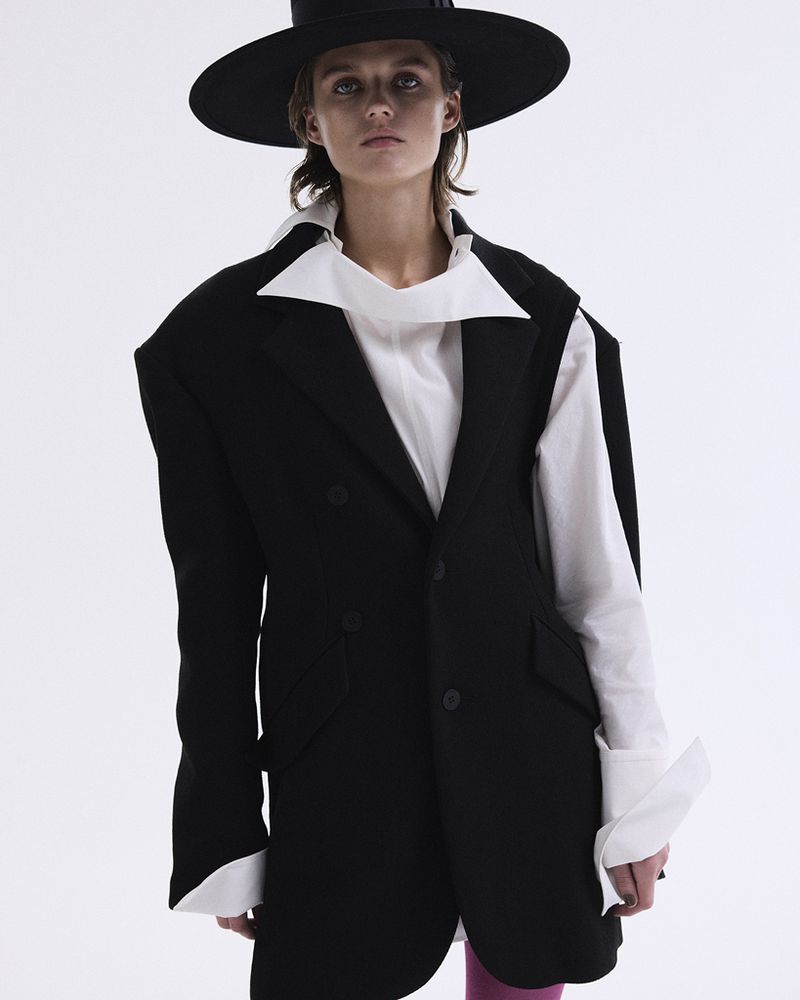 Fashion photo of a woman wearing a black blazer, wide-brimmed hat, and white blouse against a white backdrop in a studio setting.