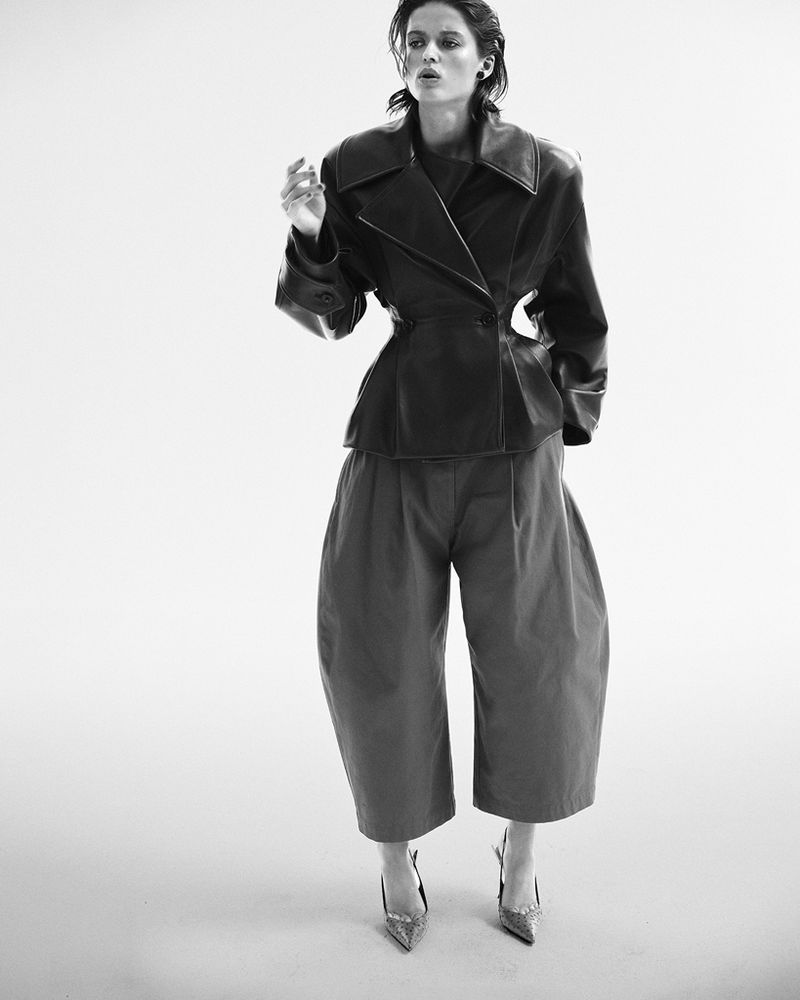 Black and white fashion portrait of a model in an oversized jacket and cropped trousers with metallic heels, posing in a studio.