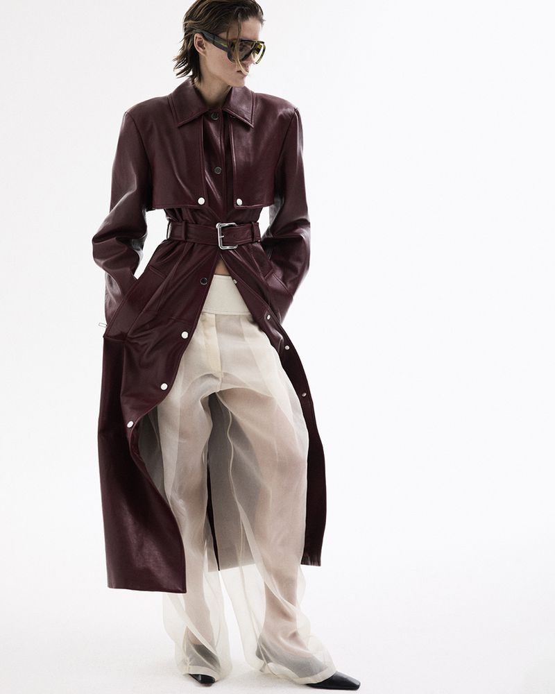 A female model poses in a studio wearing a long, red trench coat with a belt and sheer white pants, paired with dark sunglasses and black shoes.