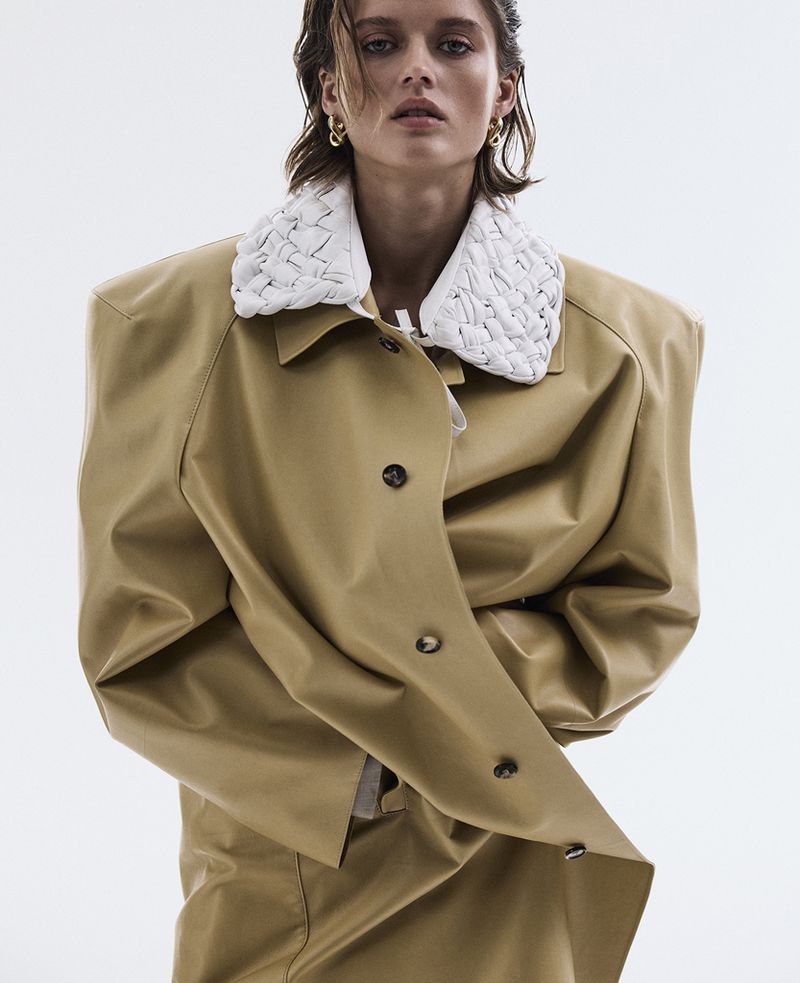 Fashion editorial shot featuring a woman in a tan coat with white braided collar against a bright white background in a studio setting.