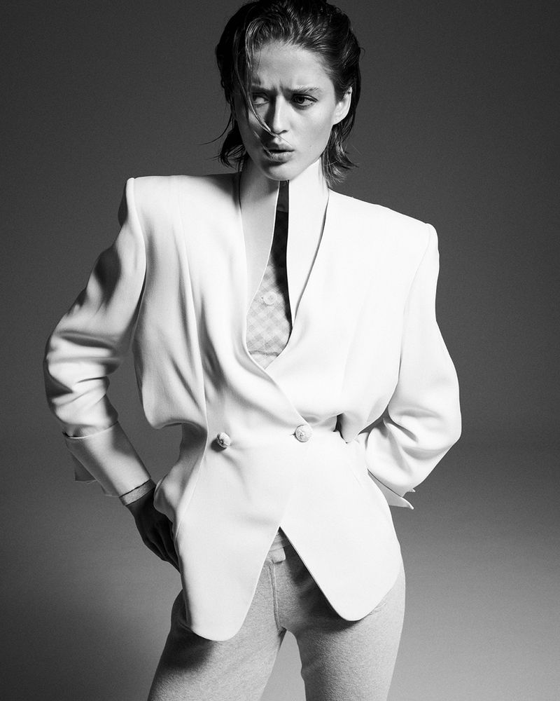 Black and white studio portrait of a woman wearing a white jacket and grey pants with her hands on her hips and looking intensely at the camera.