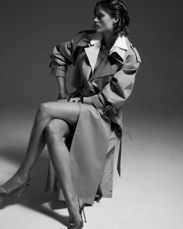 Model seated on a stool wearing a long belted trench coat and heels against a grey studio backdrop in a black and white portrait.