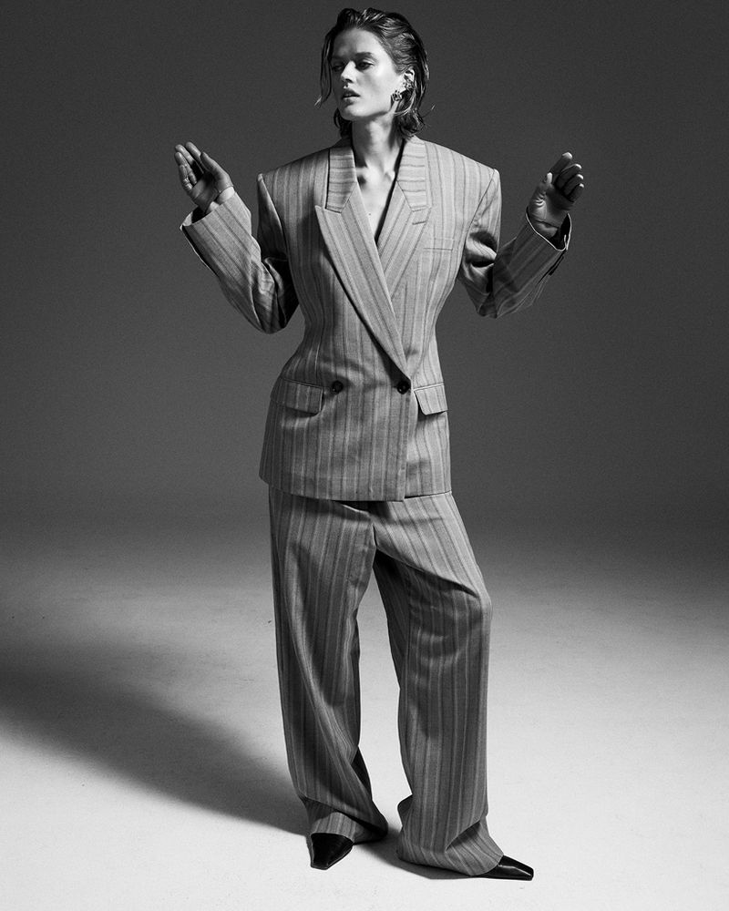 Black-and-white studio shot of a person in a pinstripe suit, hands raised near shoulders, eyes closed, wearing shiny black shoes.