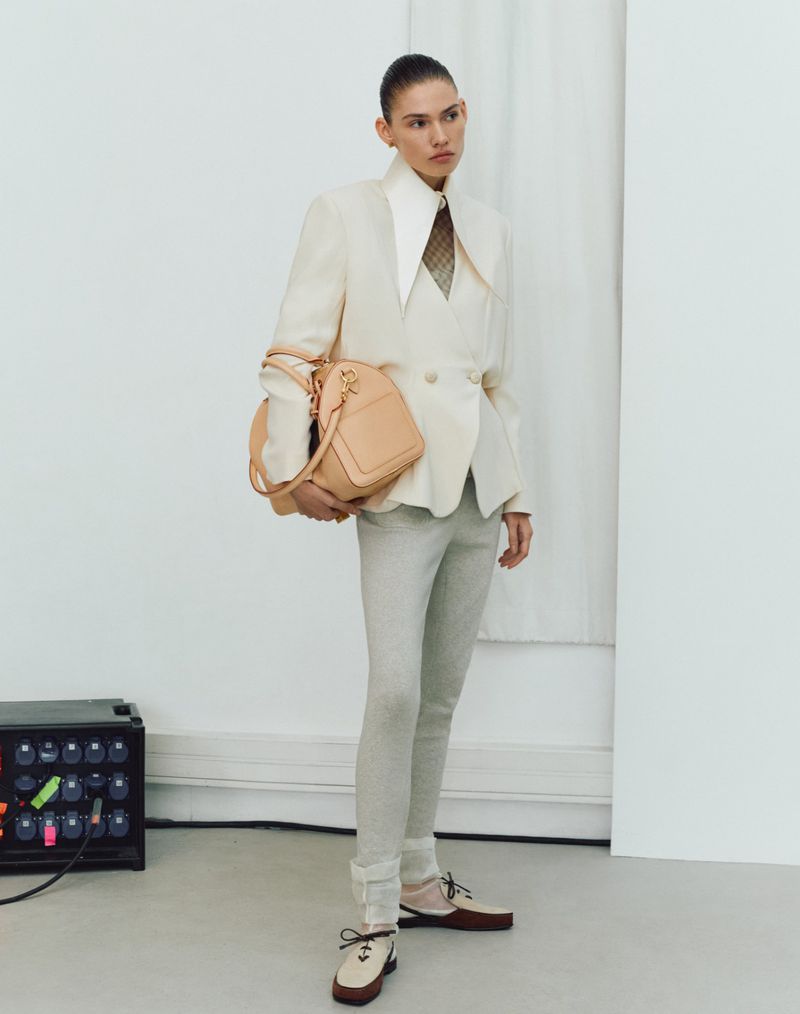 A woman in a cream blazer, light trousers, and two-tone shoes stands in a studio holding a peach-colored handbag, next to a tech device.
