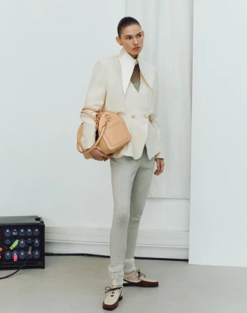 A woman in a cream blazer, light trousers, and two-tone shoes stands in a studio holding a peach-colored handbag, next to a tech device.