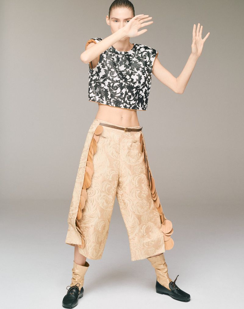 Full shot of a slender fashion model with fair skin posing in an artistic black and white top and tan textured pants in a studio.