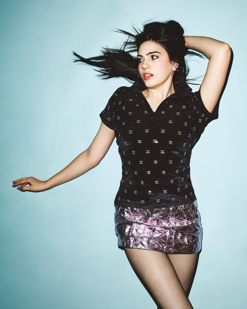 Brunette woman with hand on her head poses in black top and metallic pink miniskirt against a light blue background in studio lighting.