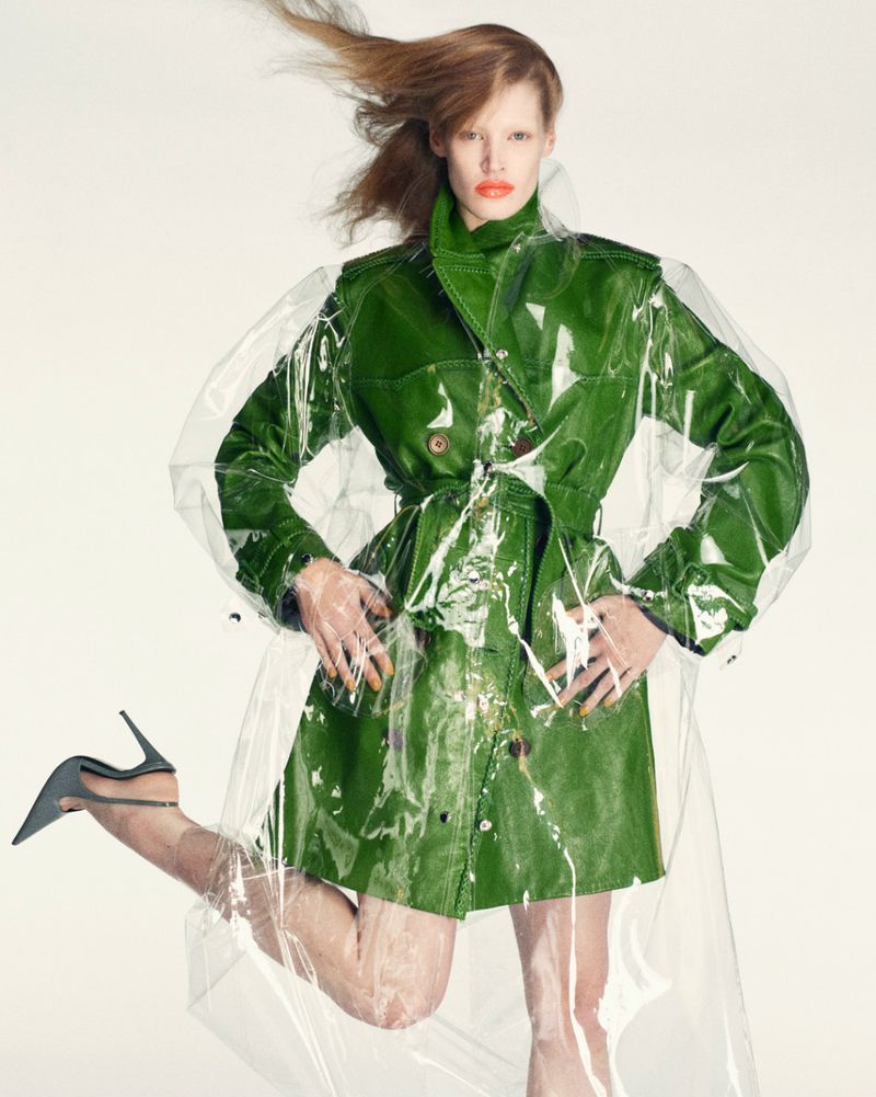 Fashionable red-haired woman poses in green trench coat with clear plastic overlay, stiletto heels against a white background.