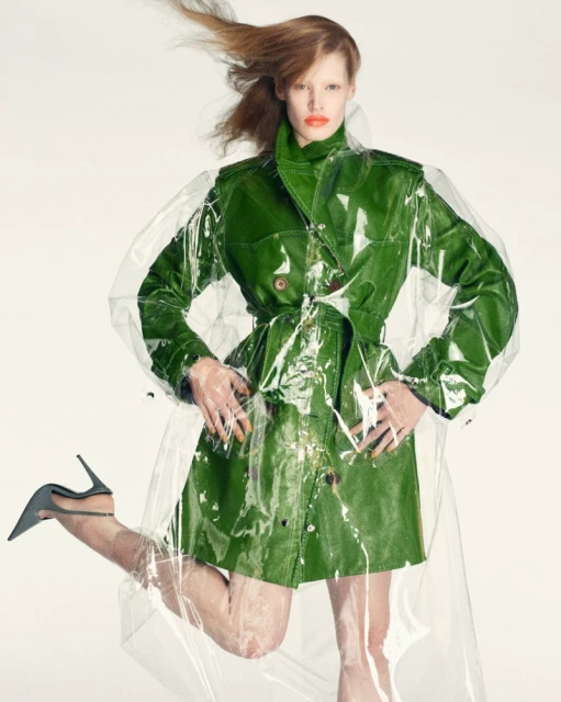 Fashionable red-haired woman poses in green trench coat with clear plastic overlay, stiletto heels against a white background.