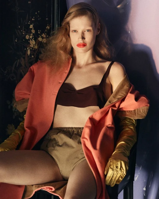 A young woman with red lipstick poses in an oversized pink coat, dark bralette, and gold gloves against a dark floral and purple background.