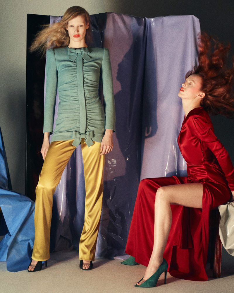 Two fashion models showcasing satin dresses and trousers in a studio setting with draped backdrop, one seated, one standing.