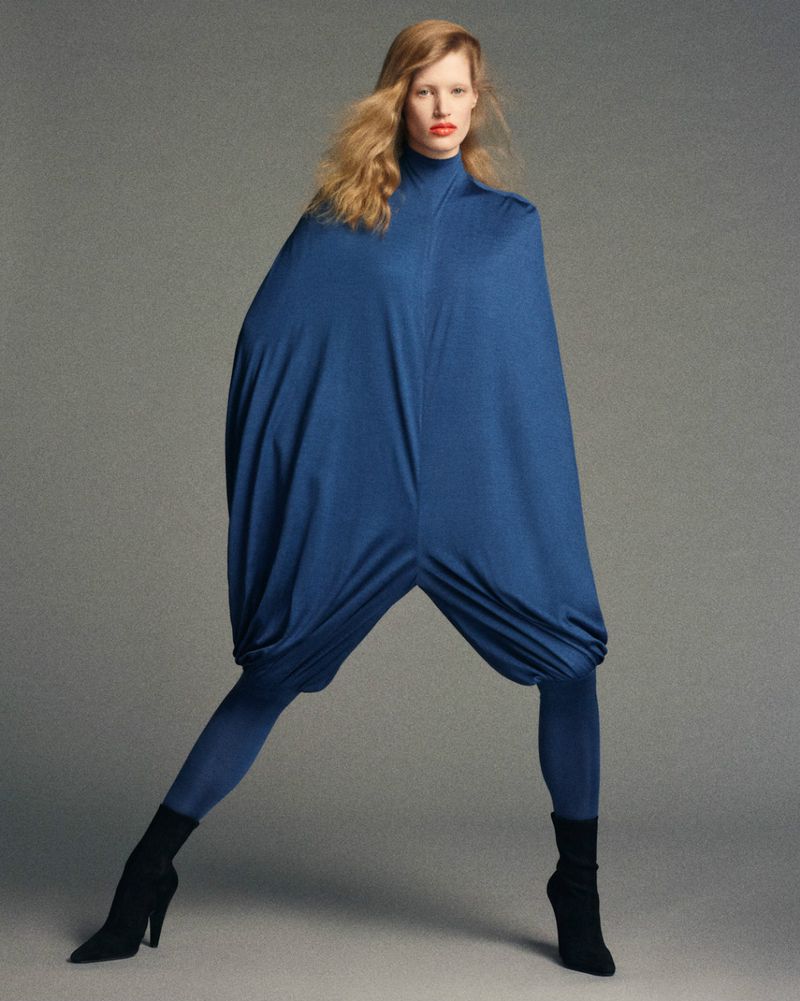 A striking fashion shot showcases a single woman in a blue, cocoon-like garment with leggings and high heels against a neutral backdrop.