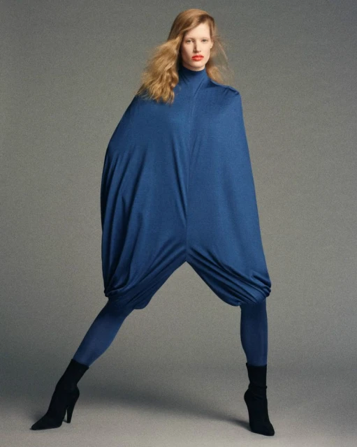 A striking fashion shot showcases a single woman in a blue, cocoon-like garment with leggings and high heels against a neutral backdrop.