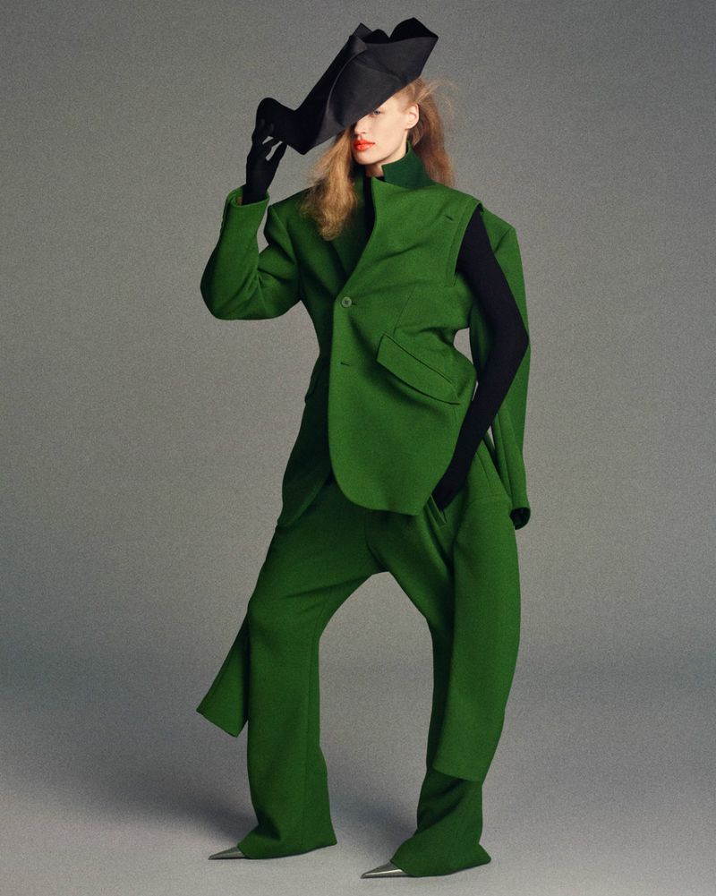 Fashionable woman in a green suit and black hat poses in a studio. The outfit features unique, oversized shapes and metallic pointed shoe tips.
