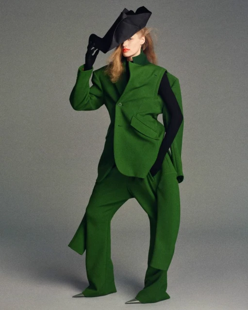 Fashionable woman in a green suit and black hat poses in a studio. The outfit features unique, oversized shapes and metallic pointed shoe tips.