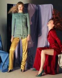 Two models in a studio setting, one wearing a green dress and gold trousers, another in a red dress with red hair whipping in motion.