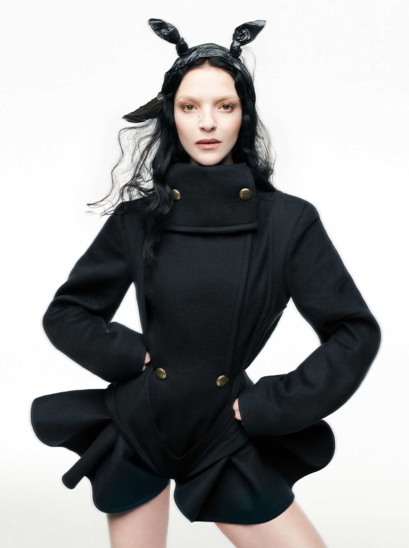 A woman with black hair, wearing a black coat, poses in a studio with a light background. She is wearing a black headpiece.