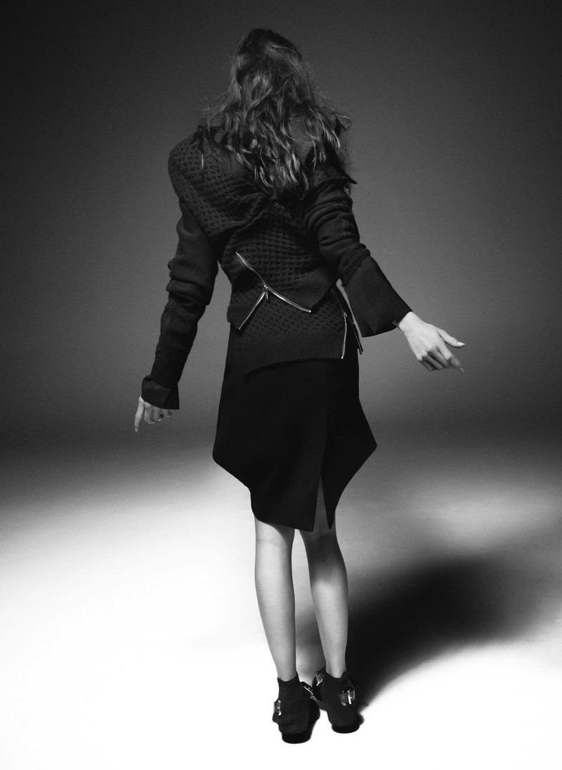 Back view of a woman wearing a black jacket and skirt in a studio shot. The background is grey, and the lighting is bright.
