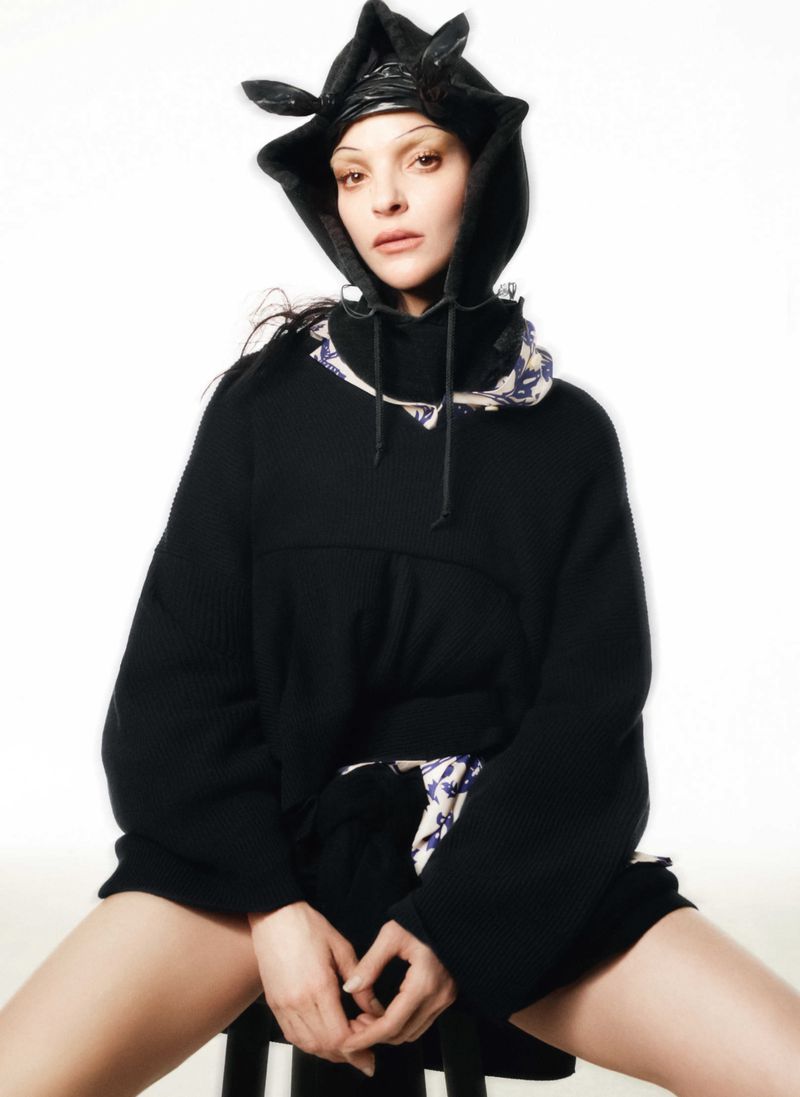 Model Mariacarla Boscono in a black hooded sweater and patterned collar against a stark white background.