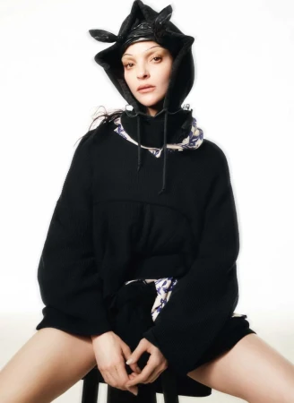 Model Mariacarla Boscono in a black hooded sweater and patterned collar against a stark white background.