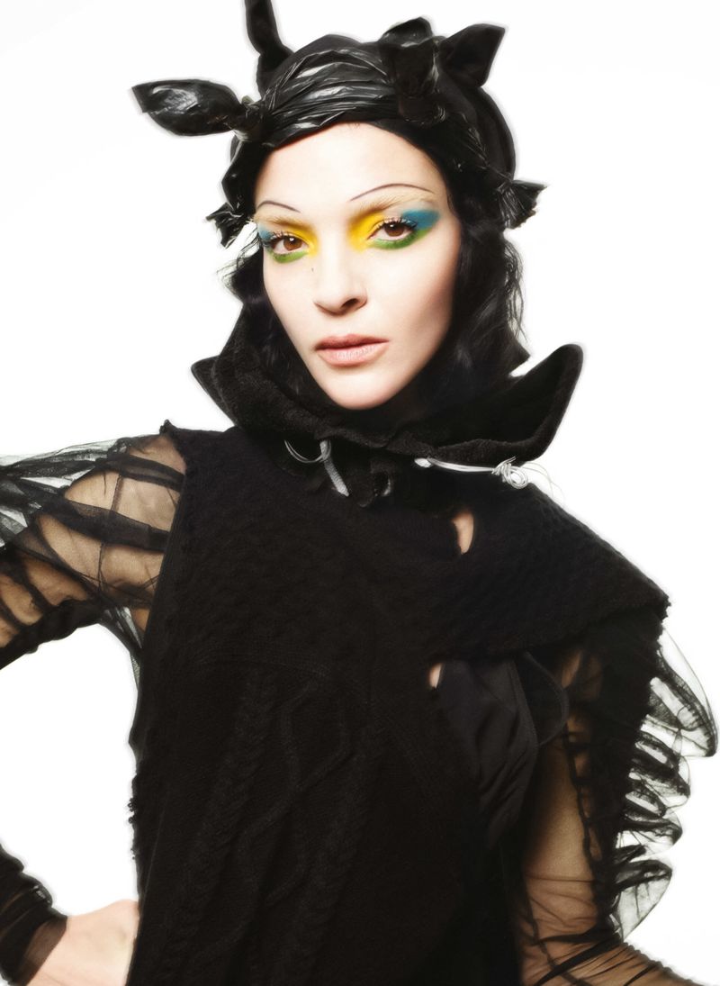 A woman poses wearing a black headdress and shoulder piece. The woman has yellow and blue makeup around her eyes. The background is white.