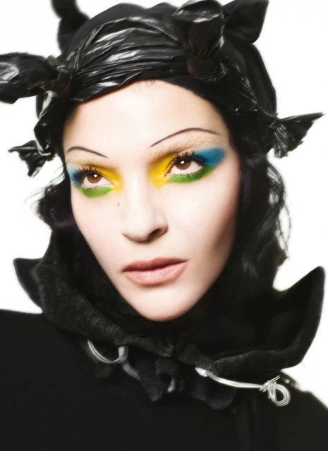 Glamorous woman wearing a black sculptural headpiece and theatrical makeup, posing in a bright studio environment. Beauty shot.