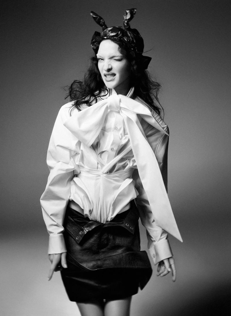 Mariacarla Boscono in a white blouse with a large bow and dark miniskirt against a seamless grey backdrop