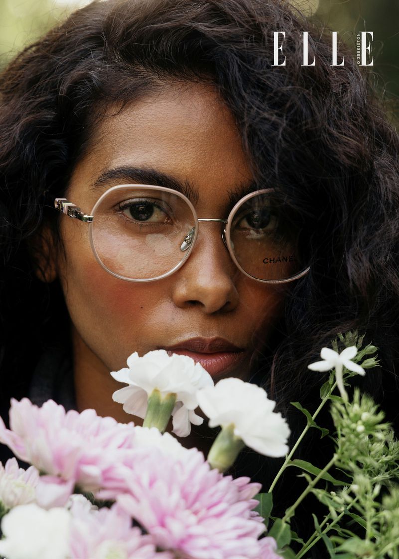 Close-up fashion shot of woman with curly hair and glasses holding white and pink flowers, wearing a dark top with a logo.