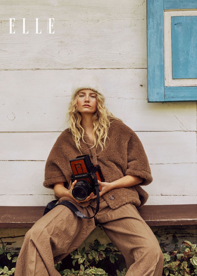 A blonde woman in brown clothing sits on a bench holding a camera in front of a white wall with a blue-framed window.