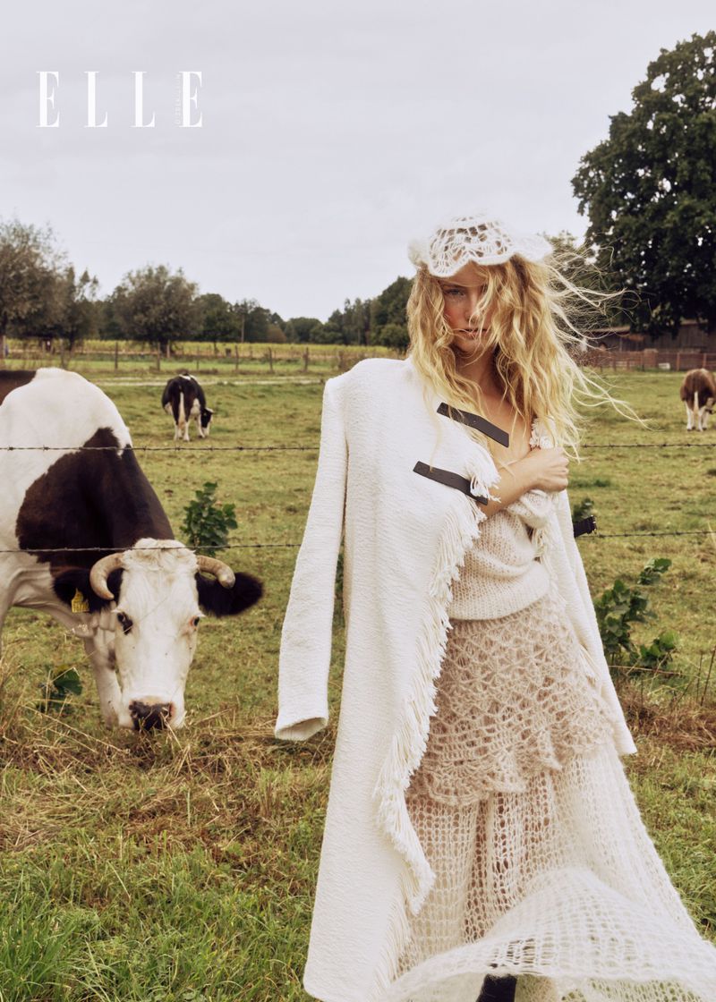 Blonde-haired woman in white lace dress and coat poses in a grassy field with black and white cows in the background; Elle magazine logo.