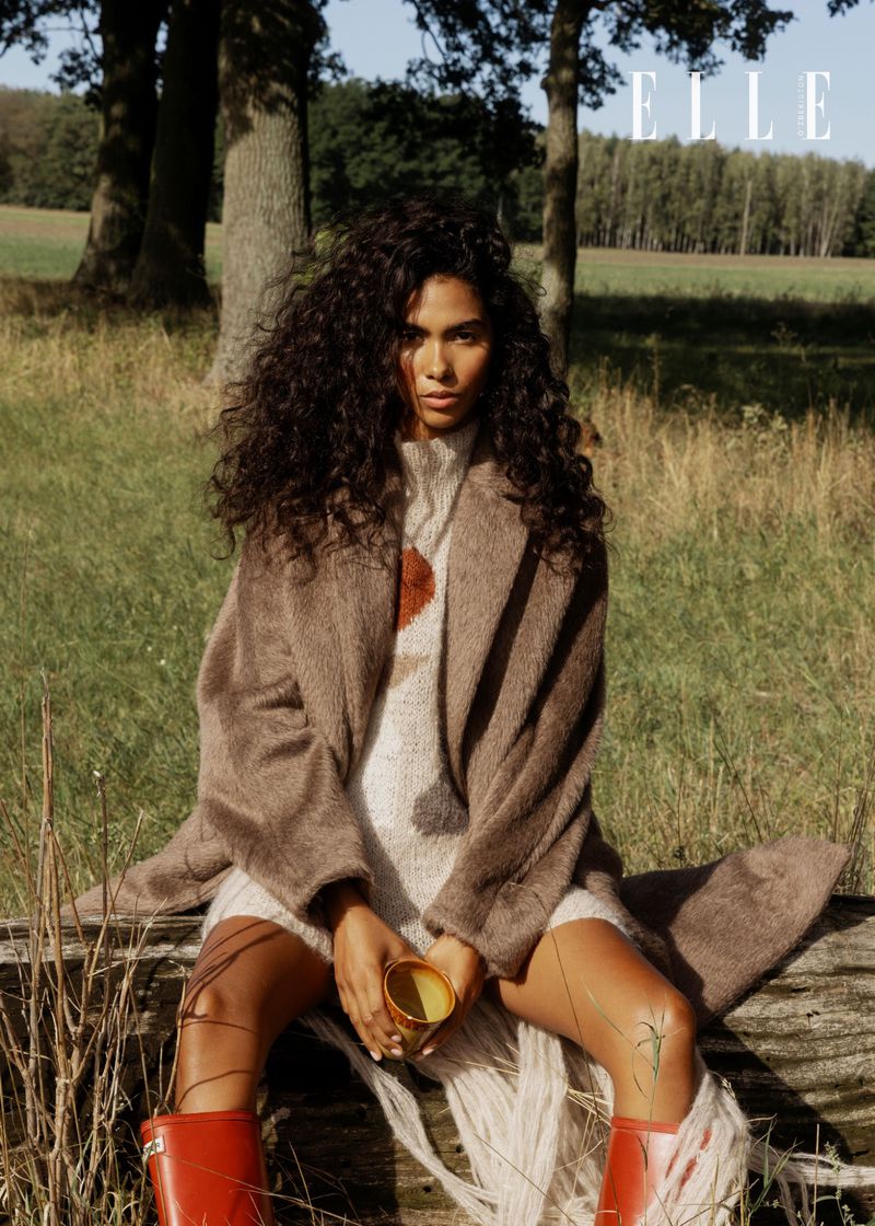 A woman with curly hair is sitting outside, wearing a sweater dress and coat, and holding a cup, with a magazine logo in the background.