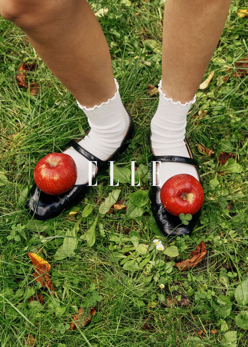Red apples on glossy black Mary Jane shoes with white ruffle socks, on green grass with the ELLE logo over the shoe straps.