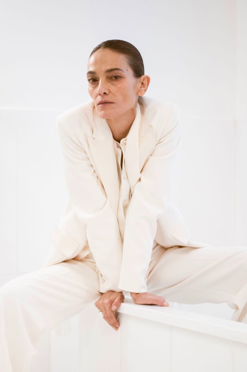 Woman in a cream-colored suit sits with bare feet in an all-white environment, posing in a relaxed manner for a fashion-forward portrait.